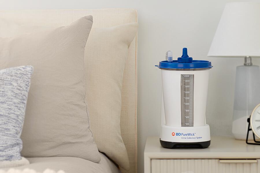 The PureWick&trade; Urine Collection System sits on a nightstand beside a bed and lamp
